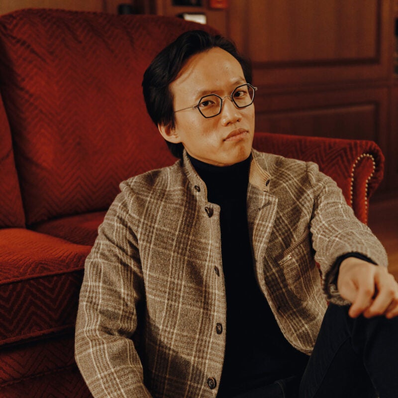 A man in a light brown plaid jacket sits in front of a rusty red couch