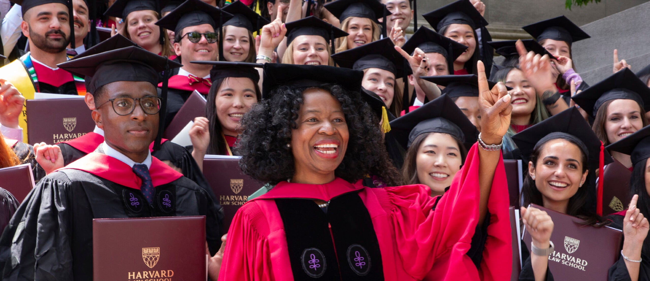 A group of graduates in cap and gown pose for a photo with their professor gesturing a one with her finger.