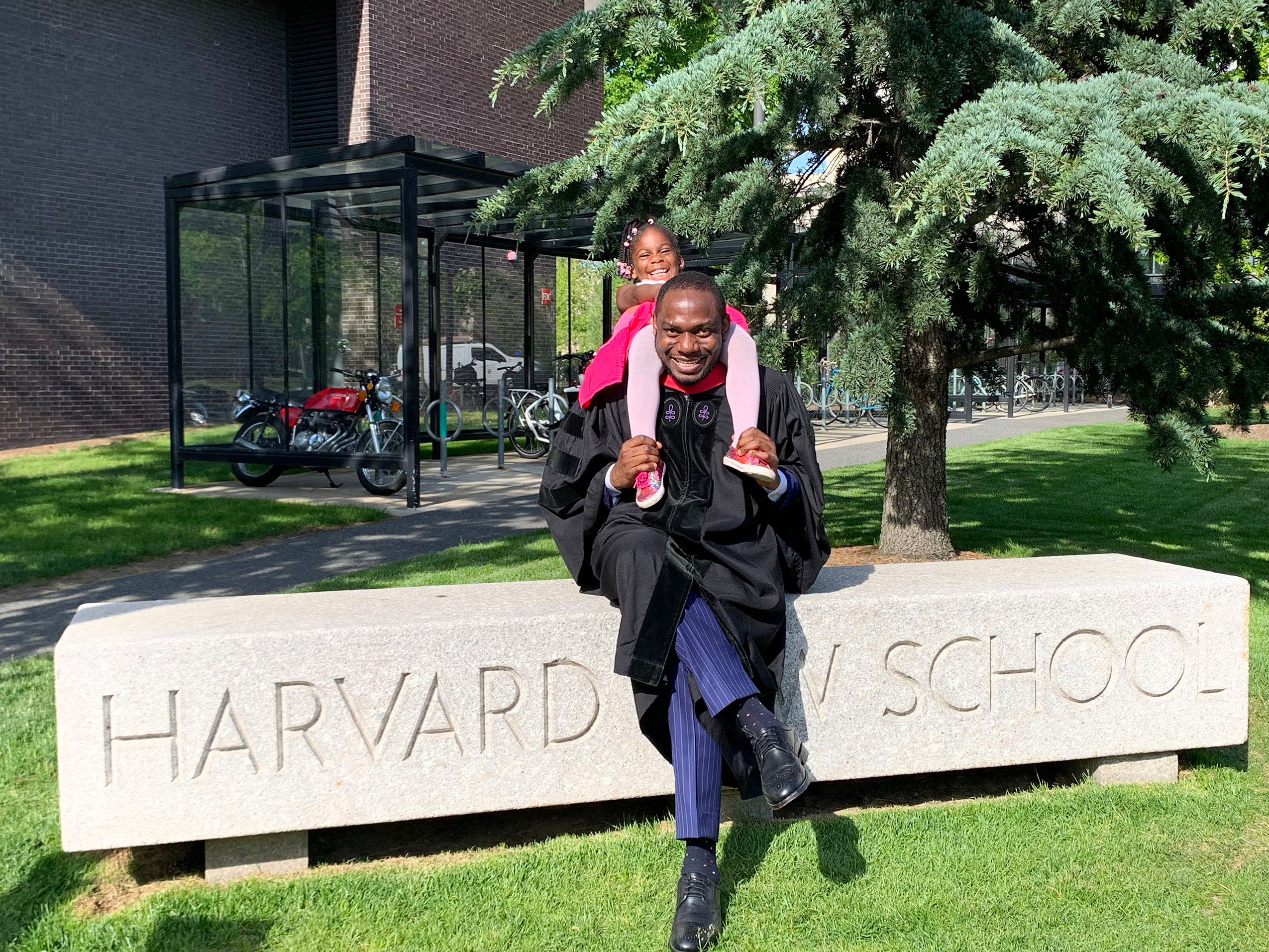 A man in a graduation gown with his young daughter on his shoulders, laughing, sits outside on a stone bench that says Harvard Law School.