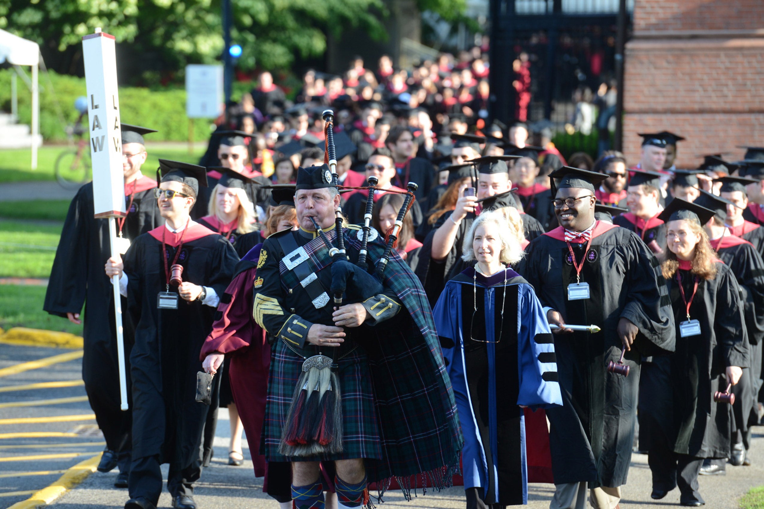 Parade of graduates in robes lead by a bagpiper in a green plaid.
