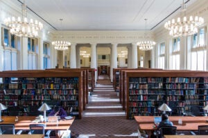 Wide view of the reading room where students study at long tables. The room features large overhead lighting, many bookshelves, pillars, and latin inscriptions on the walls.