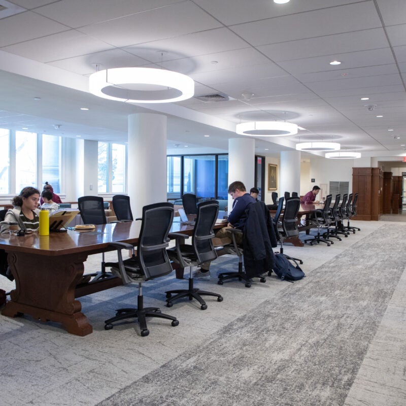 A large open space with overhead lighting shows people at long desks doing work withe armchairs and bar seting on the edges.