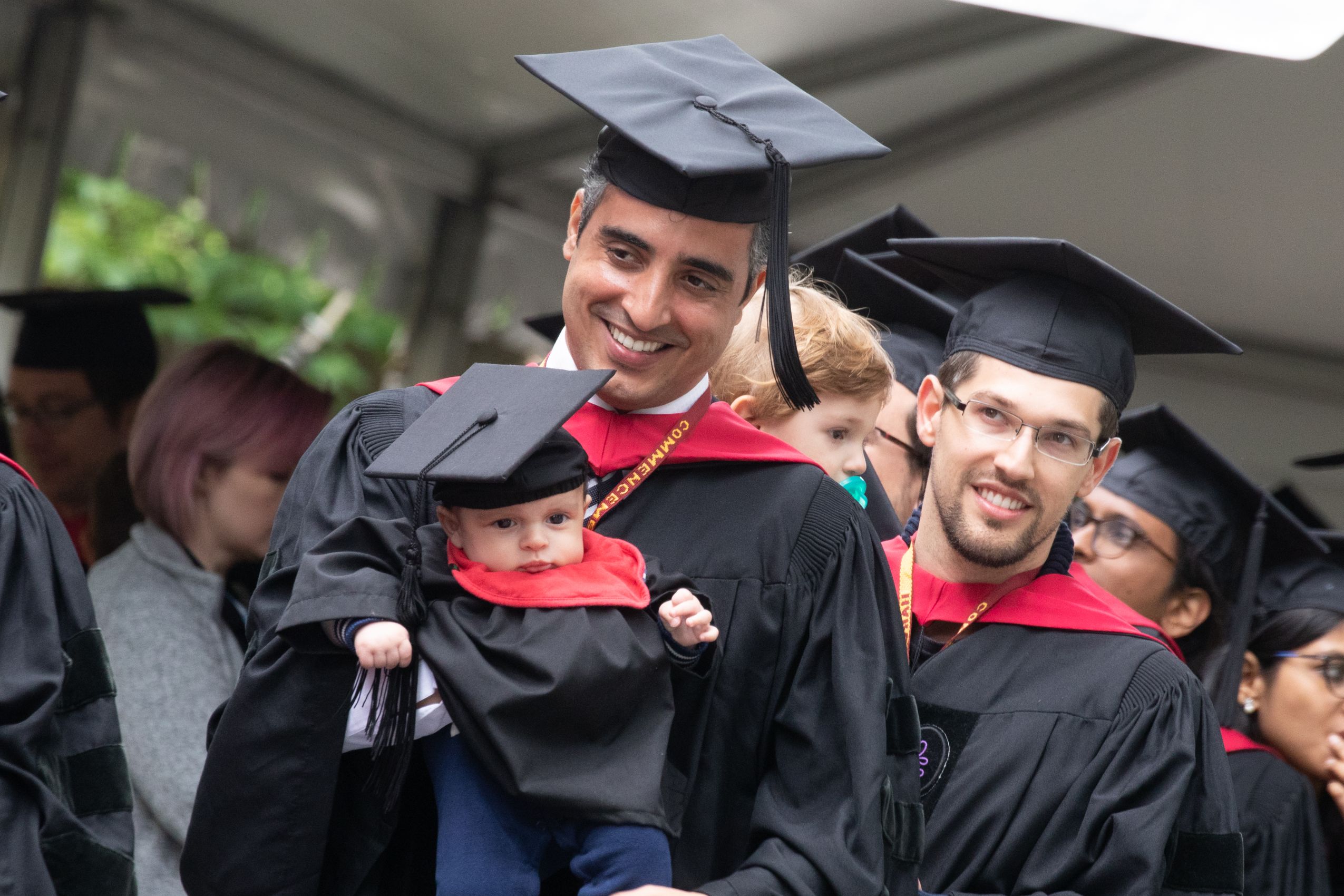 Man wearing Commencement gown and hat holding a baby wearing a commencement gown and hat
