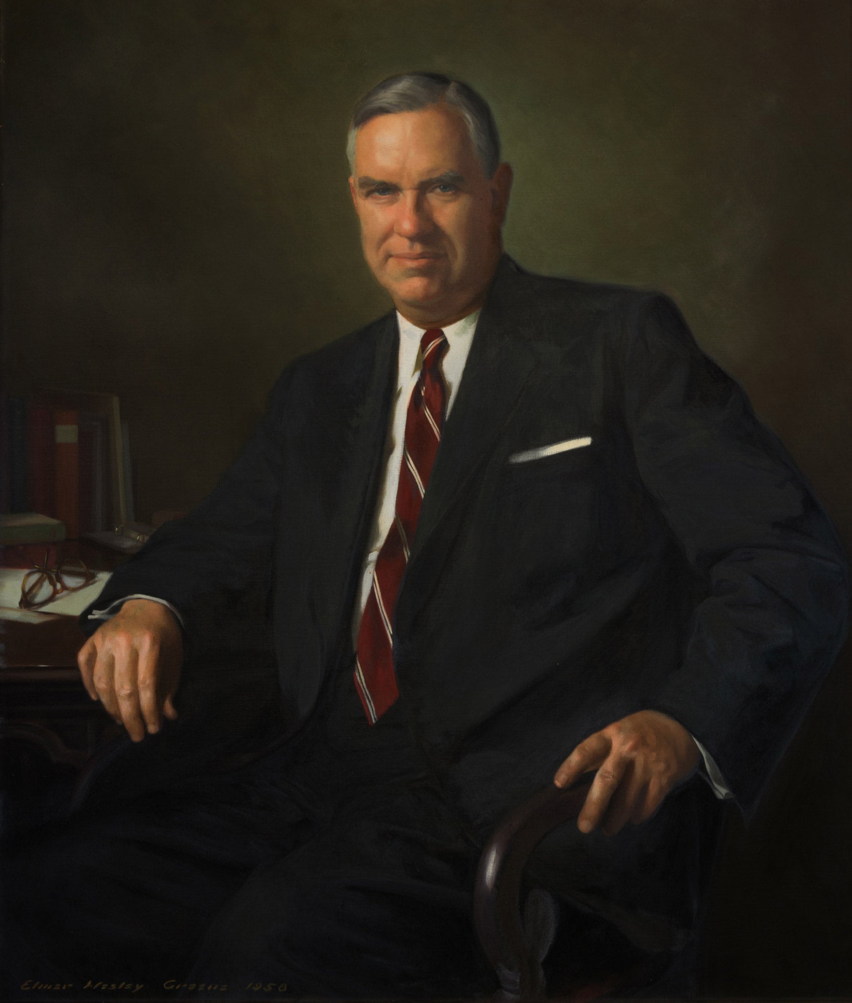 A man in a suit sitting in a chair with an elbow leaning on a table and facing forward