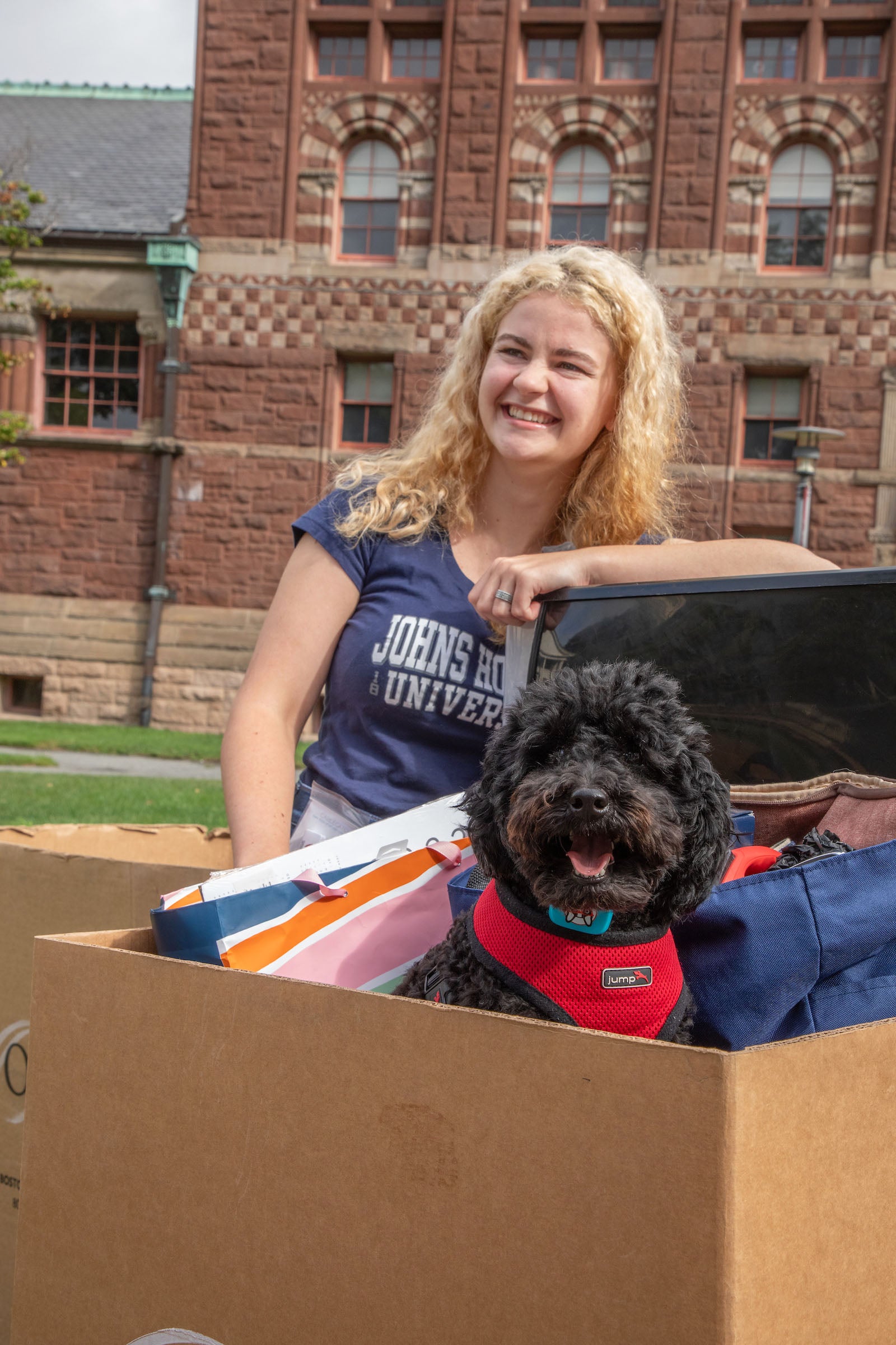 A student stands with a moving box outside, her black, fluffy dog sits in the box.
