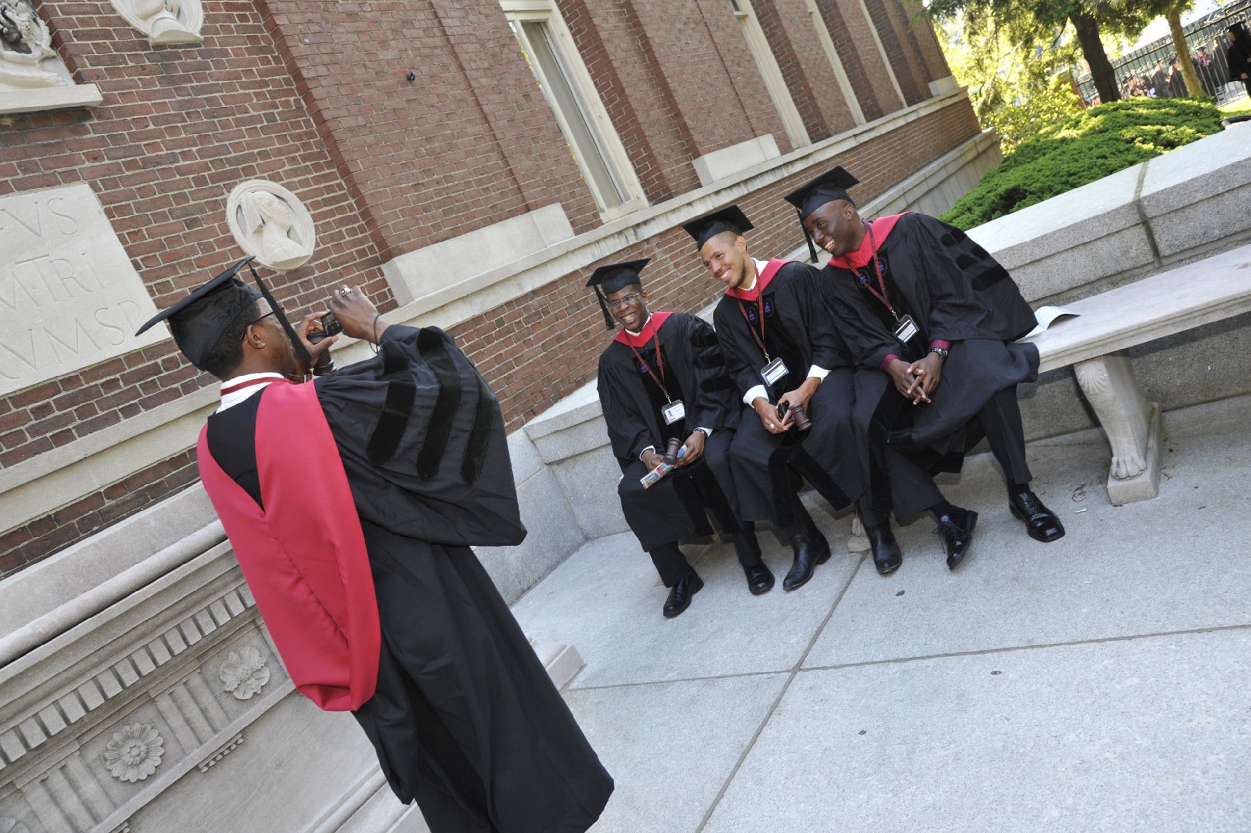 Graduate in black Commencement robe with red hood takes a cell phone photo of three classmates sitting on a bench