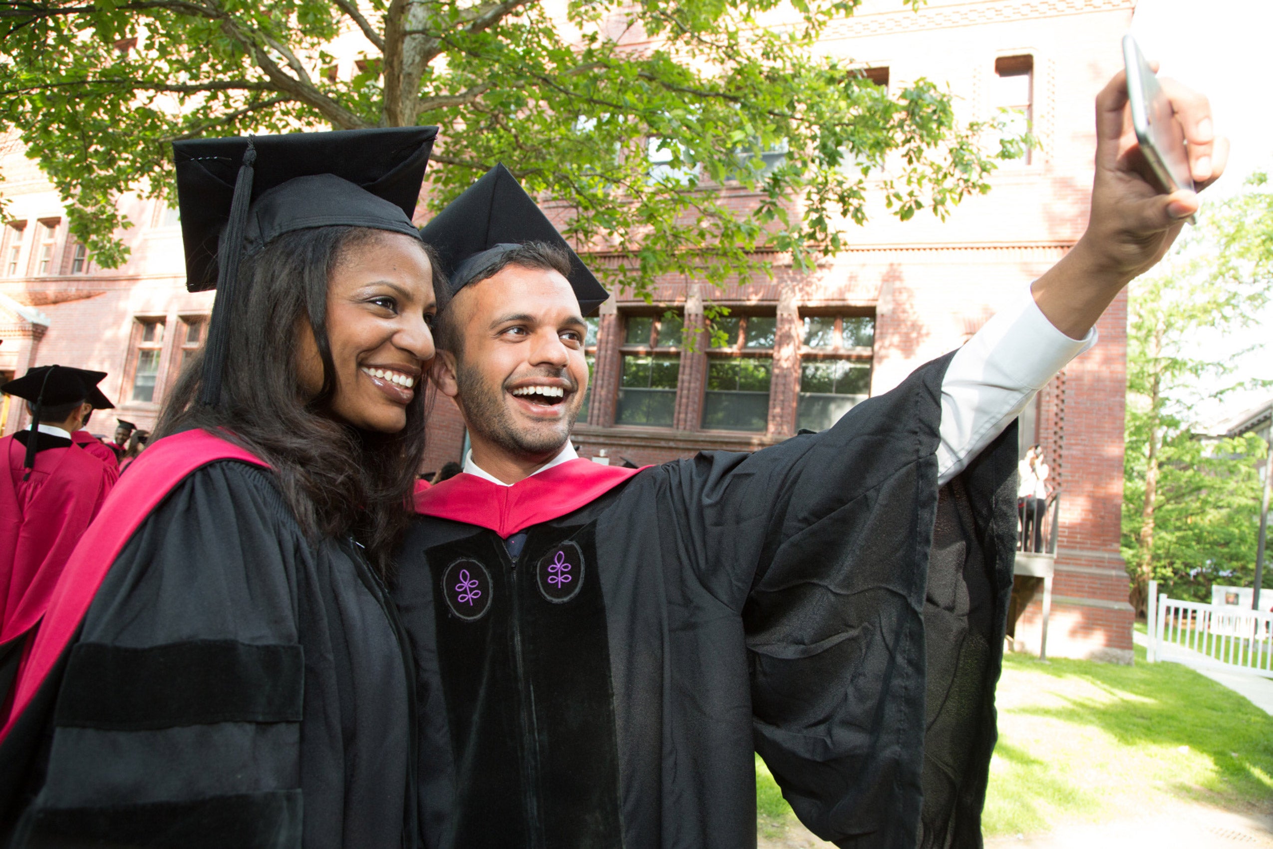 A man and a woman wearing black Commencement robes take a selfie near Harvard Yard