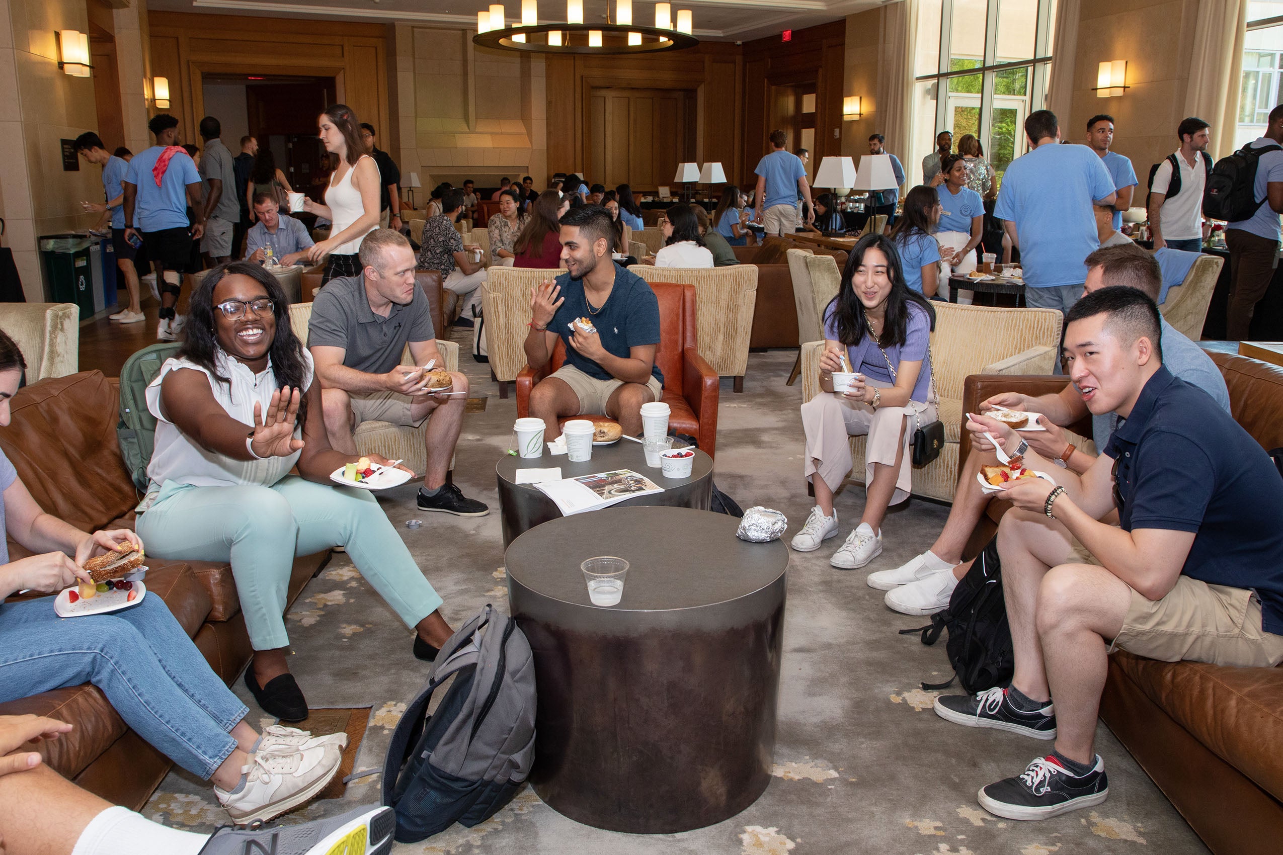 A group of studentes sitting inside on couches and chairs, eating and talking.