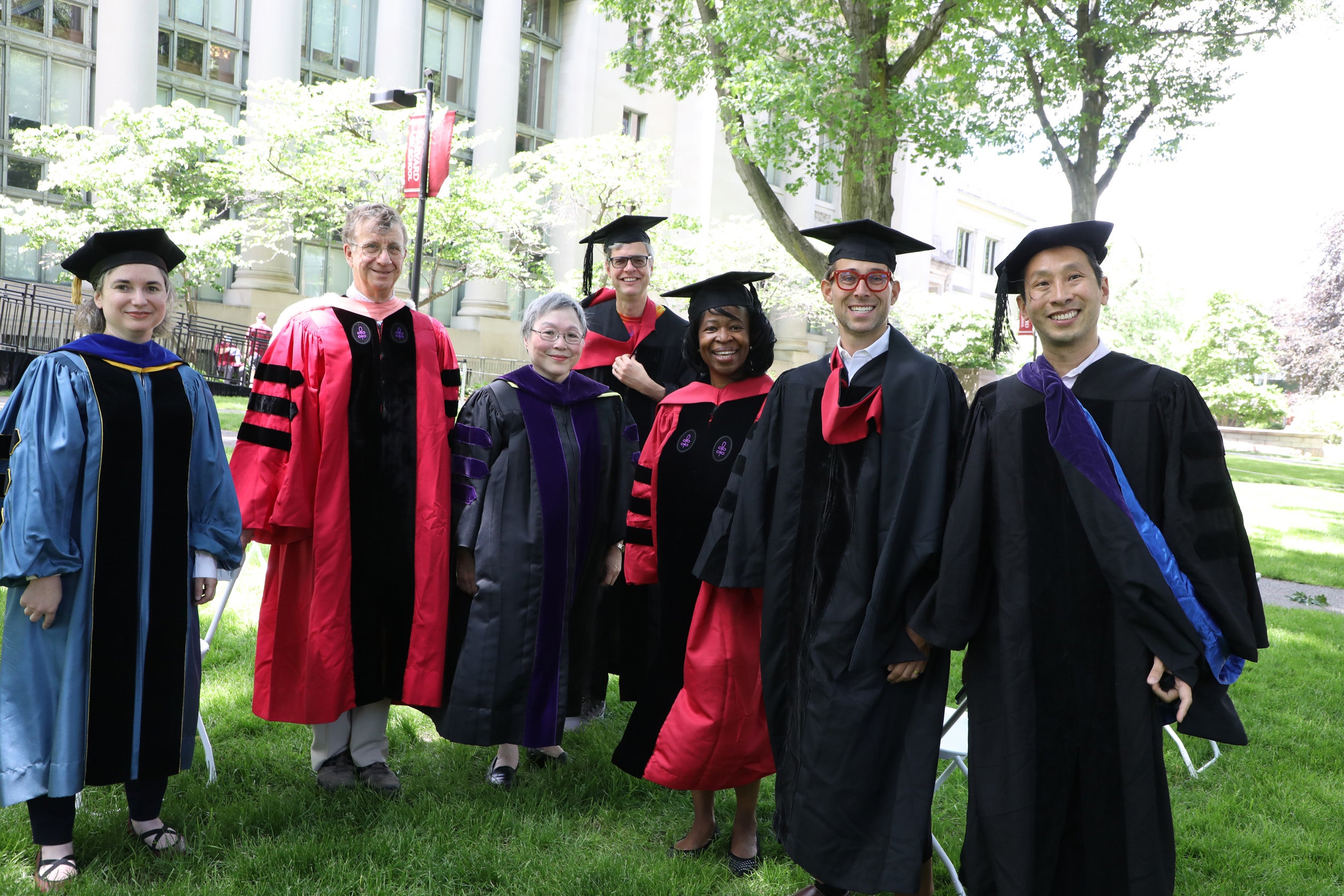 Men and women faculty pose on the lawn in front of a building wearing in red or black commencement attire