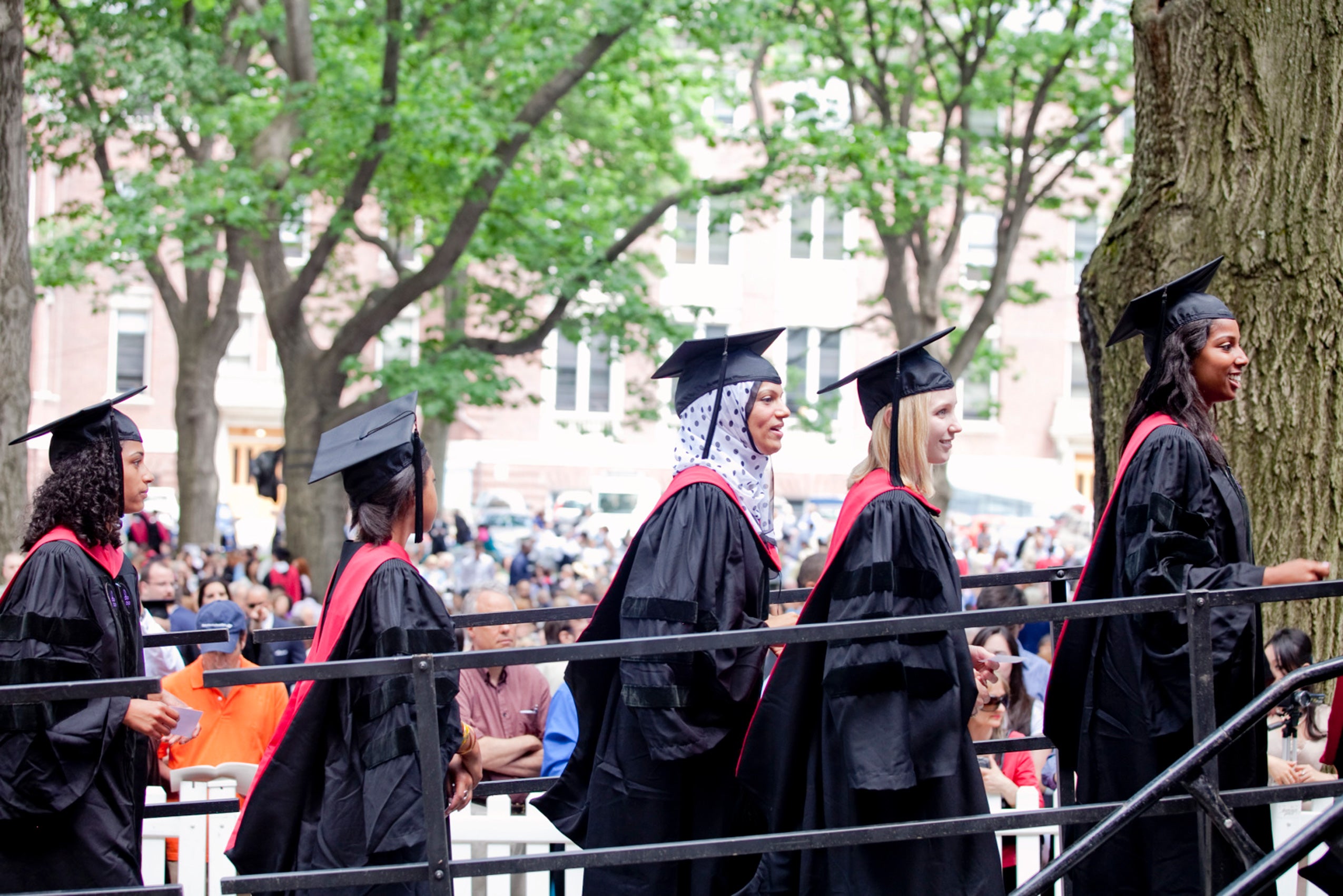 Six graduates in black robes and red hood walk up a ramp to a stage in front of a field filled with people