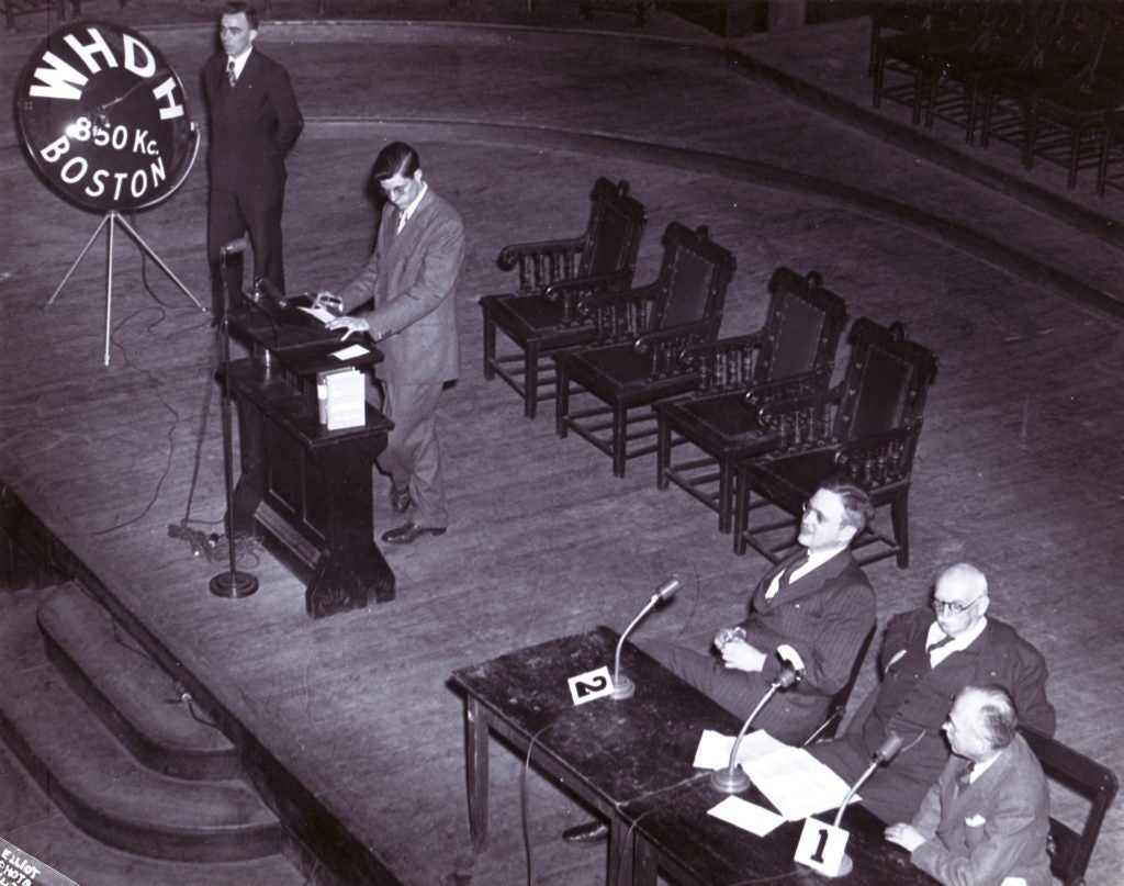 A view from above of a stage with a man speaking at a podium near a sign that says WHDH Boston and three men sitting at a table nearby with