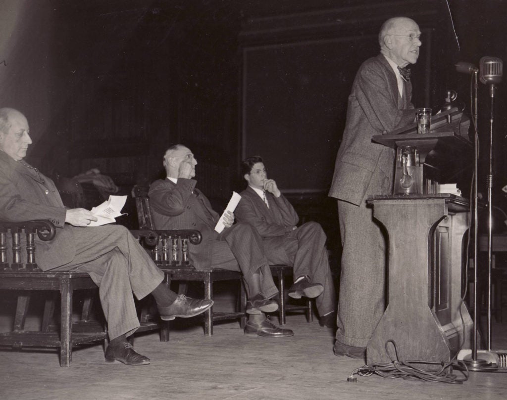 A man leaning on the podium speaking and two older men and one younger man sitting in chairs behind him