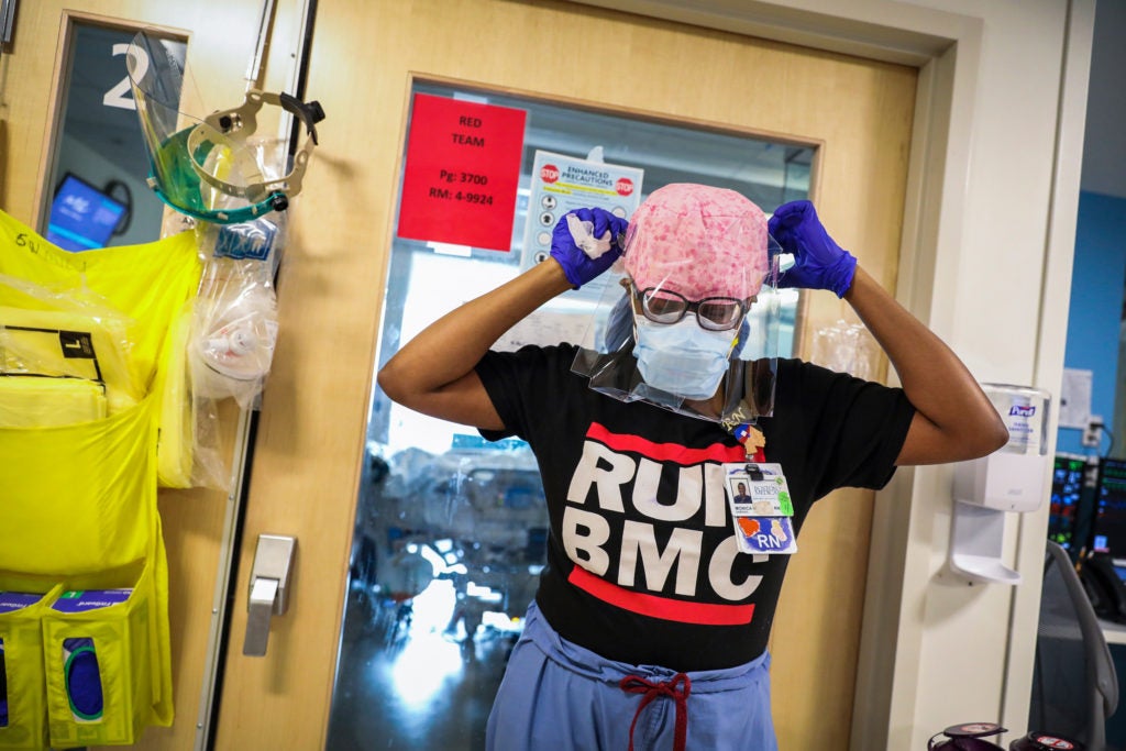 A nurse wearing a protective head cap and black shirt that says BMC removes her face shield as she stands outside of a room in a hospital