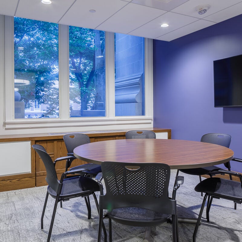 Private room with big glass windows. In the center is a round table with six chairs and flat screen on the wall for projection.