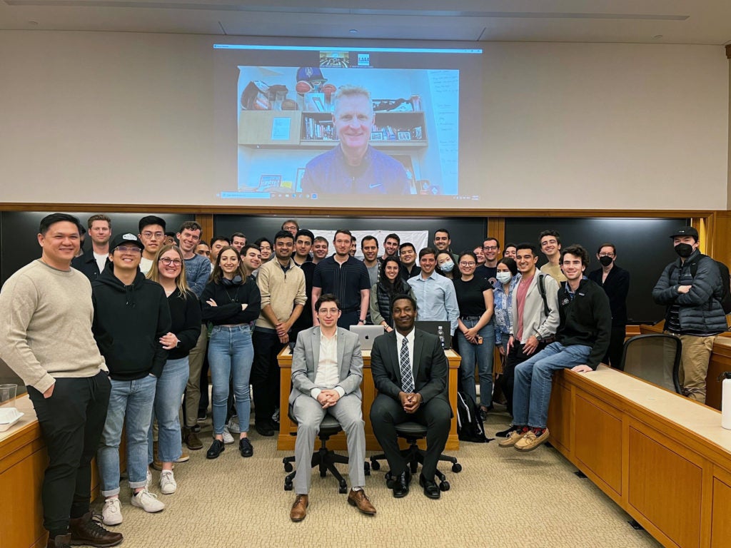 A large group of students stands for a group photo in a classroom below a video screen with Steve Kerr