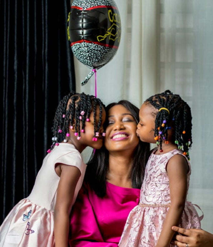Onyi Iyizoba with her young daughters kissing her cheaks and a helium balloon in the background