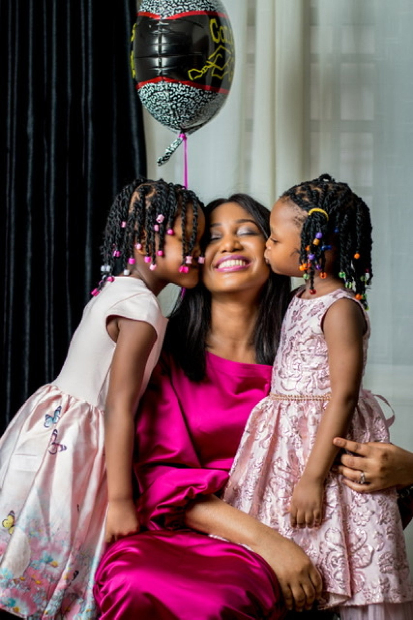 Woman in a pink silk dress with a young children pink dresses on either side kissing her cheek and balloon with the word congratulations above.above her.