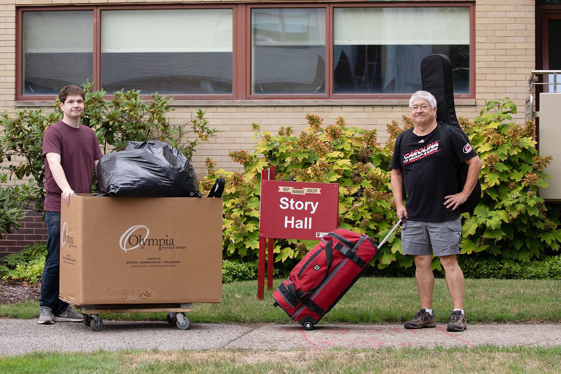 A man and his son standing outside a dorm with a suitcase and moving box.