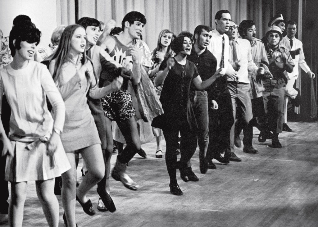 A black and white photo of two lines of people singing and dancing on a stage