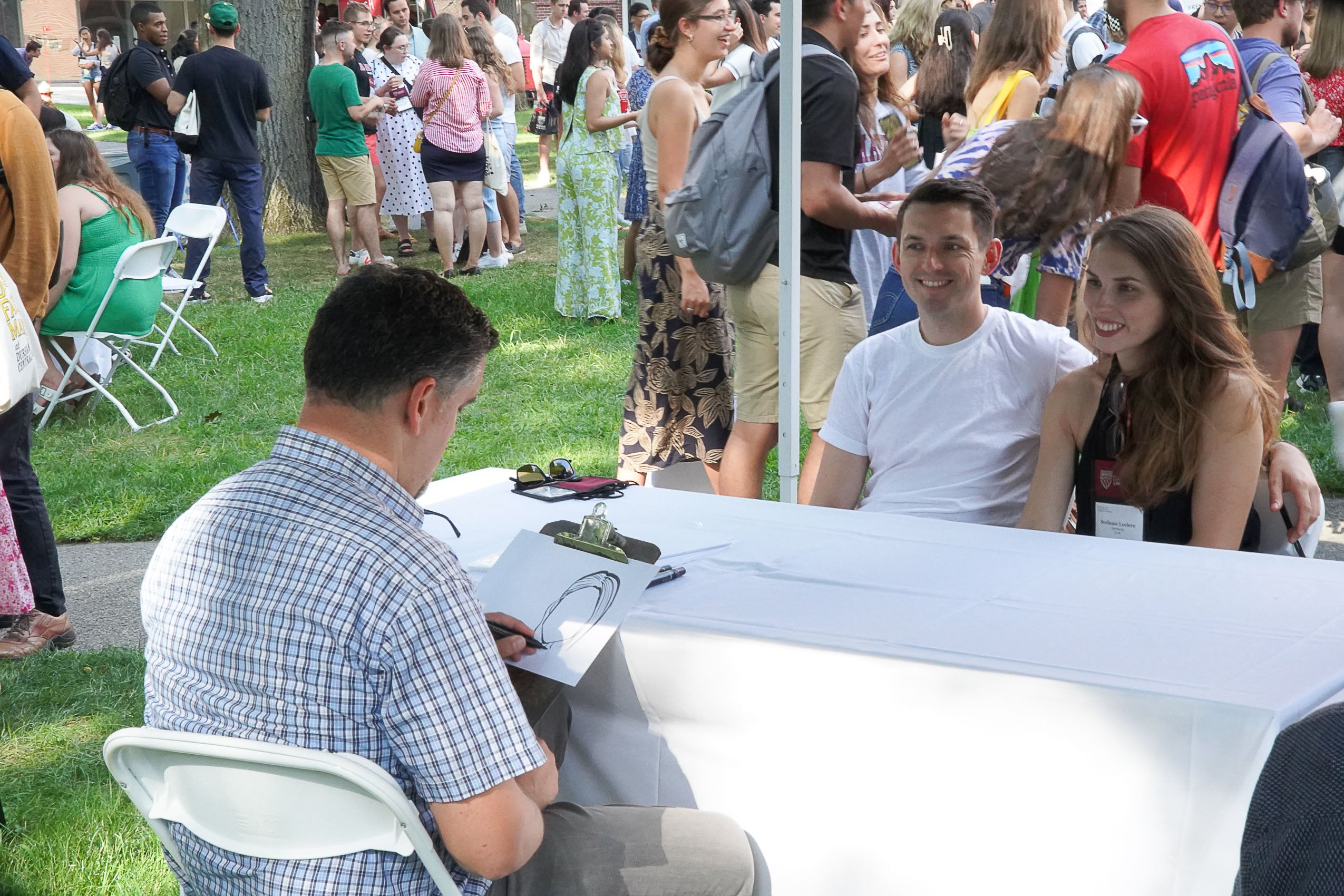 A man and woman sitting outside at a table while a caricature artist draws them.