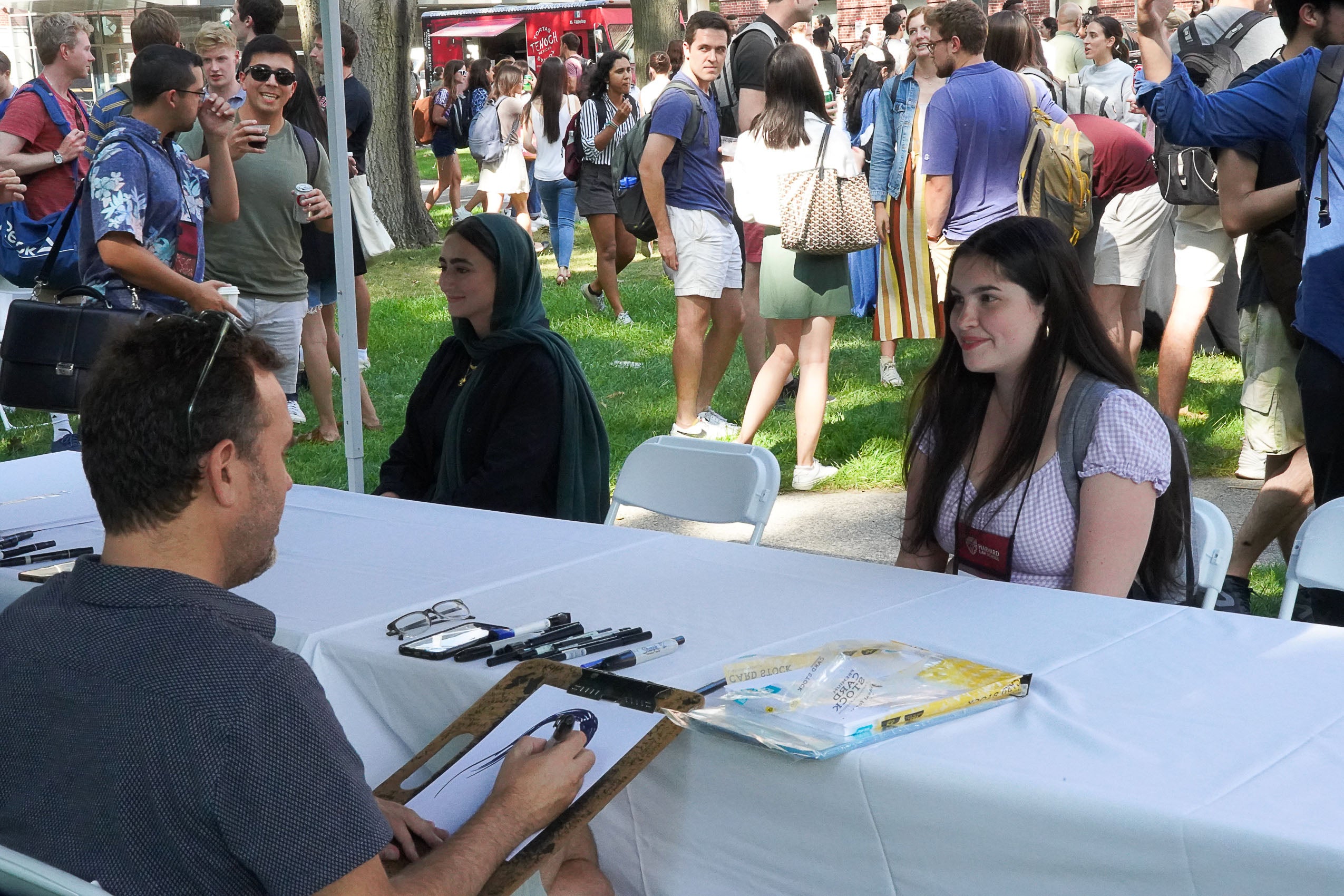 Two women sitting outside at a table while caricature artists draws them.
