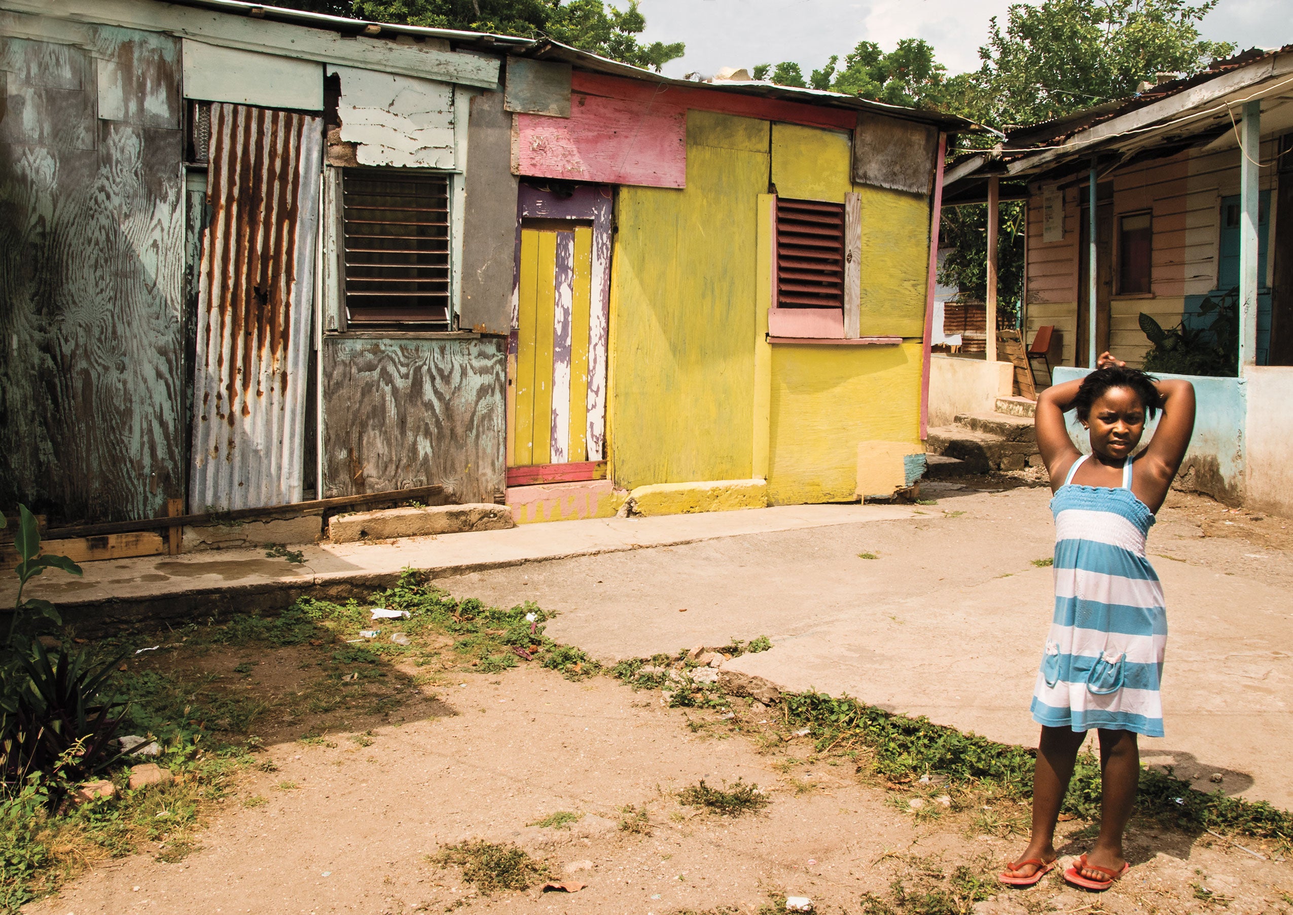 a young girl in a striped dress stands in front of a building made out of scraps of wood and metal