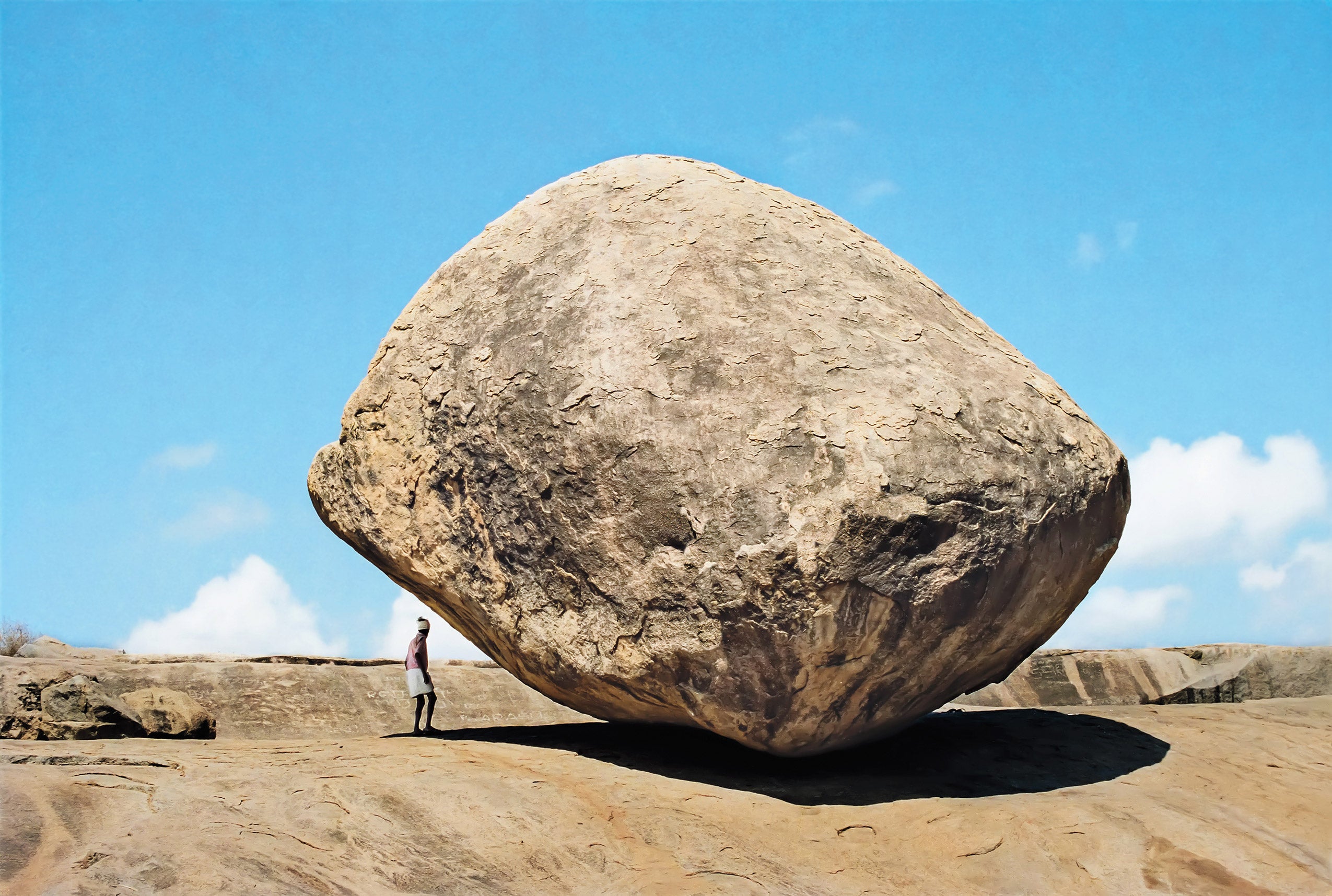 A man stands beneath to a large rock