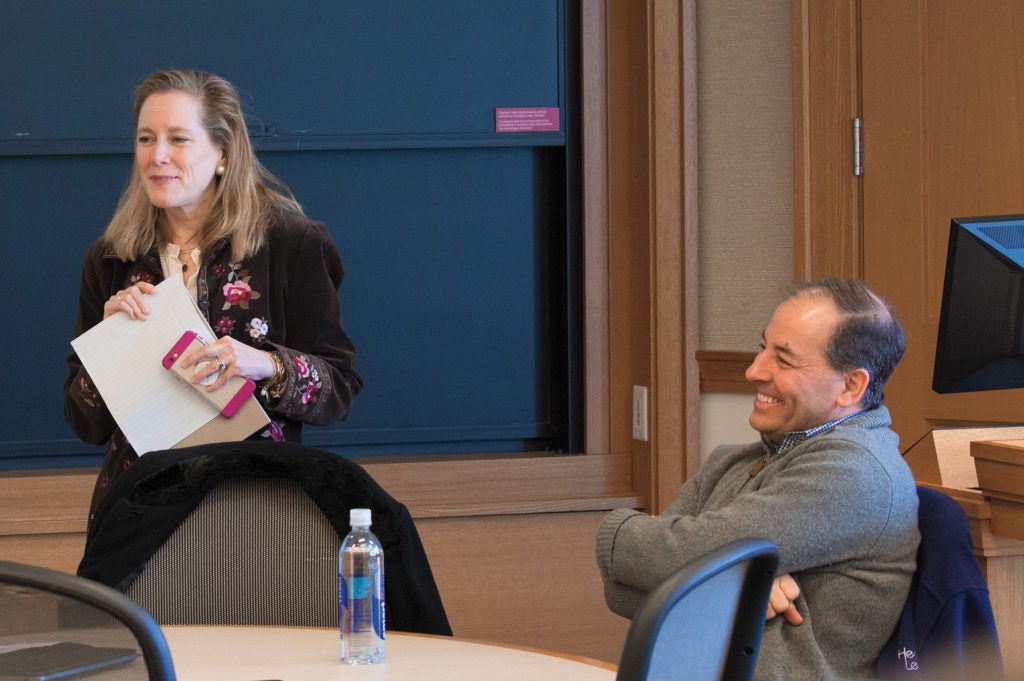 A woman standing at the front of a classroom and a man to her left sitting at a table nearby