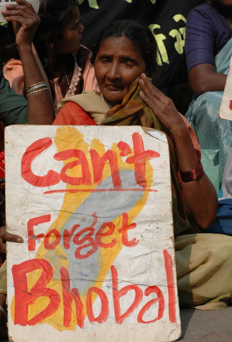 A woman seated with a sign in front of her that reads Can't Forget Bhopal.