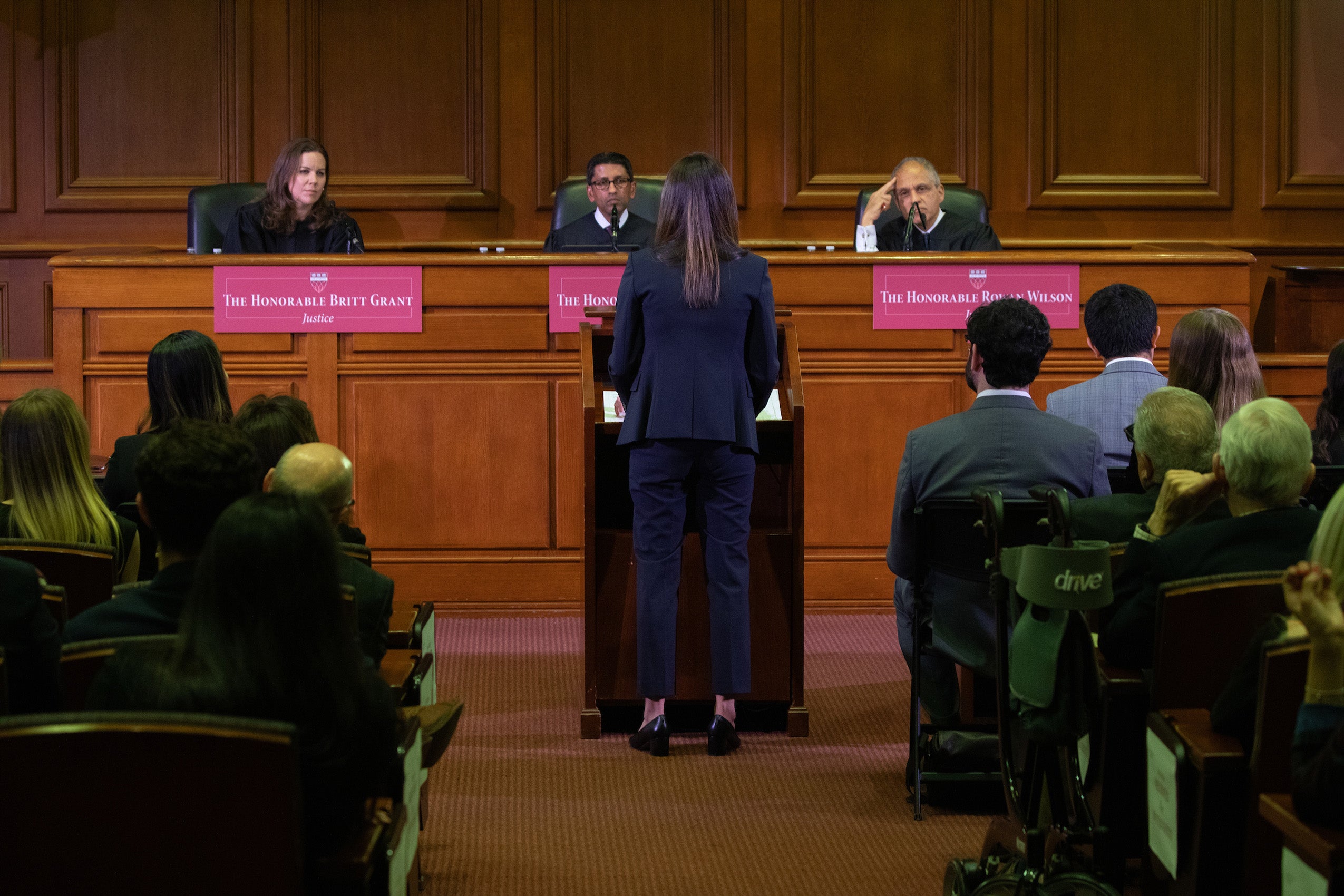 A woman stands at a podium as three judges sitting on a panel in front of her listen.