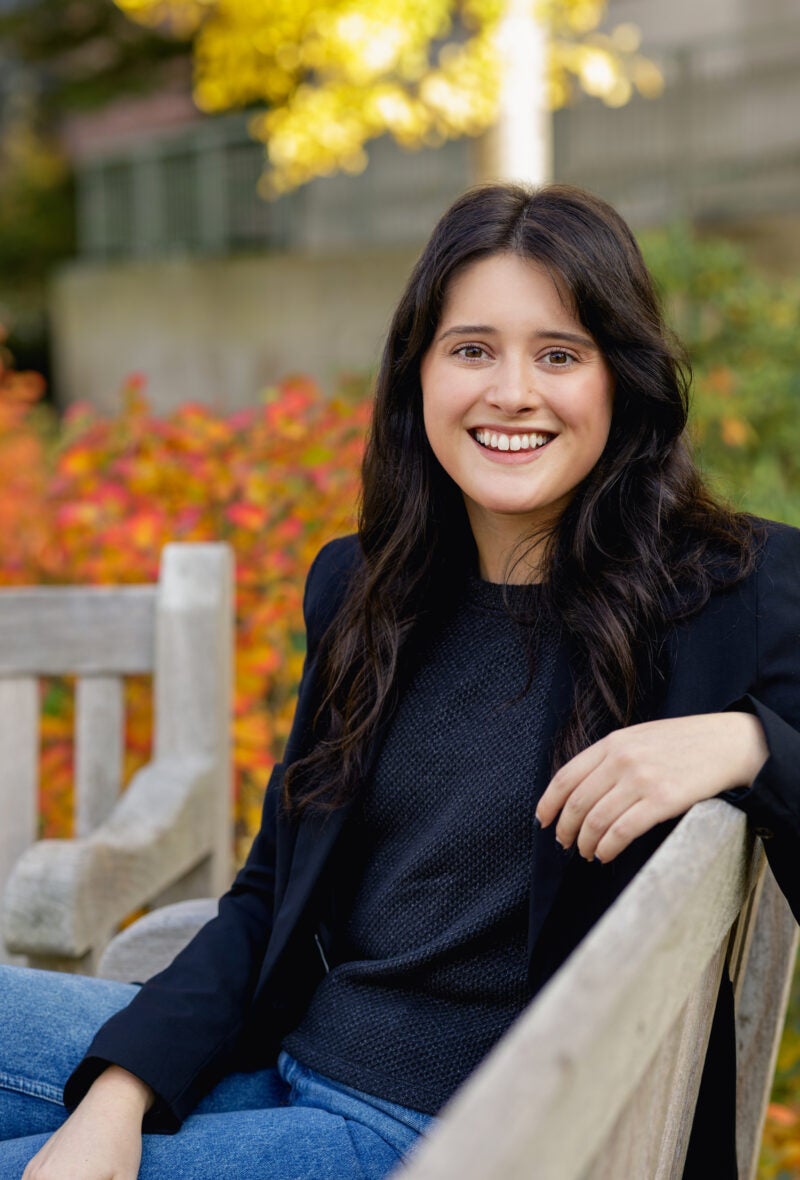 A woman in a black blazer sits on a bench on the Harvard Law School campus.