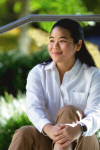 A woman in a white shirt sits in front of trees on the Harvard Law School campus.