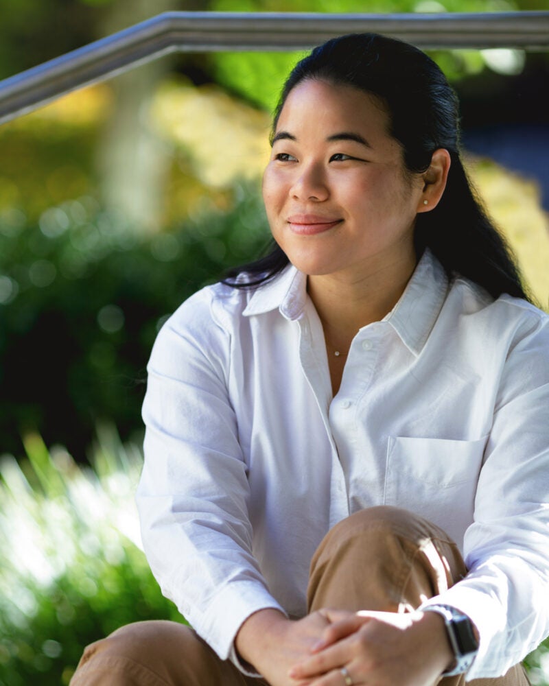 A woman in a white shirt sits in front of trees on the Harvard Law School campus.