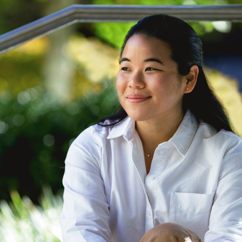 A woman in a white shirt sits in front of trees on the Harvard Law School campus.