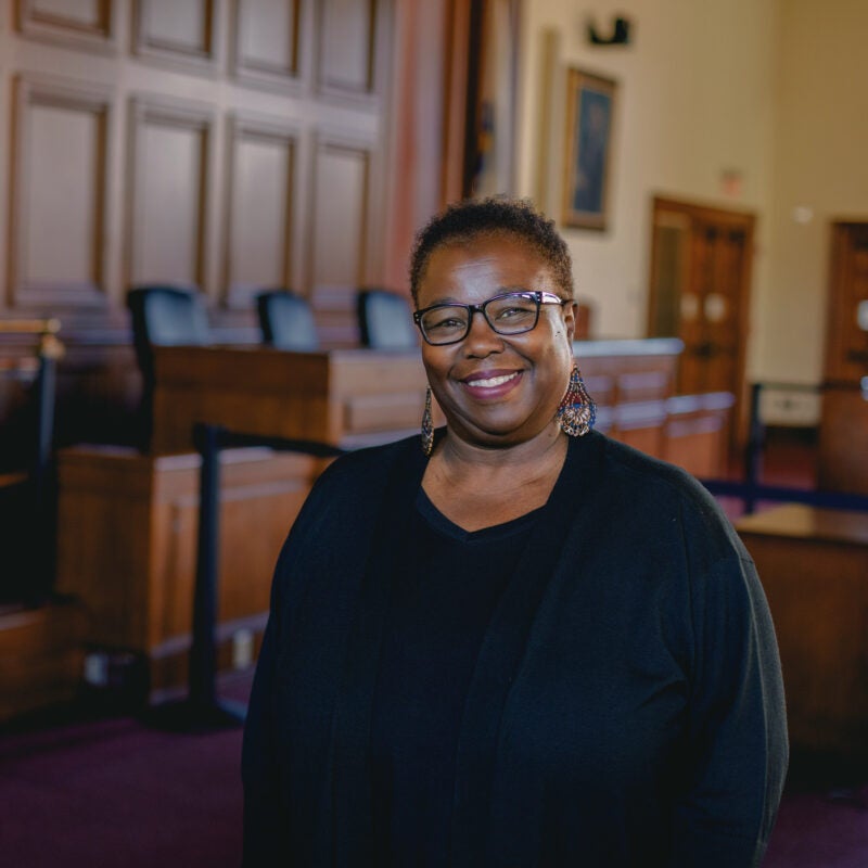 A woman in a black sweater poses in front of judges bench in the Ames Courtroom at Harvard Law School.