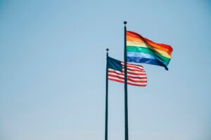 An US flag and a rainbow flag are on two poles next to each other blowing in the wind.