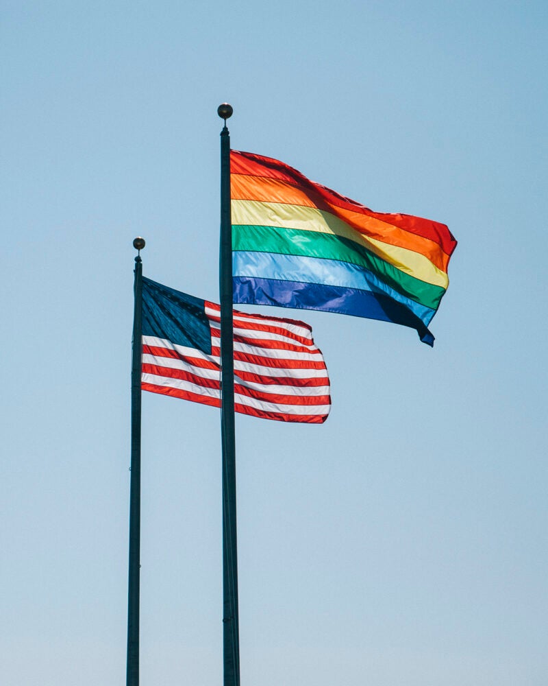 An US flag and a rainbow flag are on two poles next to each other blowing in the wind.