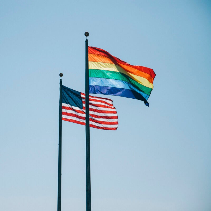 An US flag and a rainbow flag are on two poles next to each other blowing in the wind.