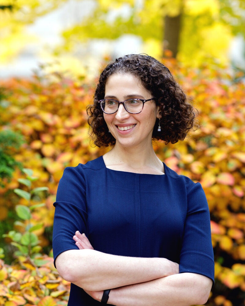 A woman in a blue dress poses in front of fall leaves on the Harvard Law School campus.