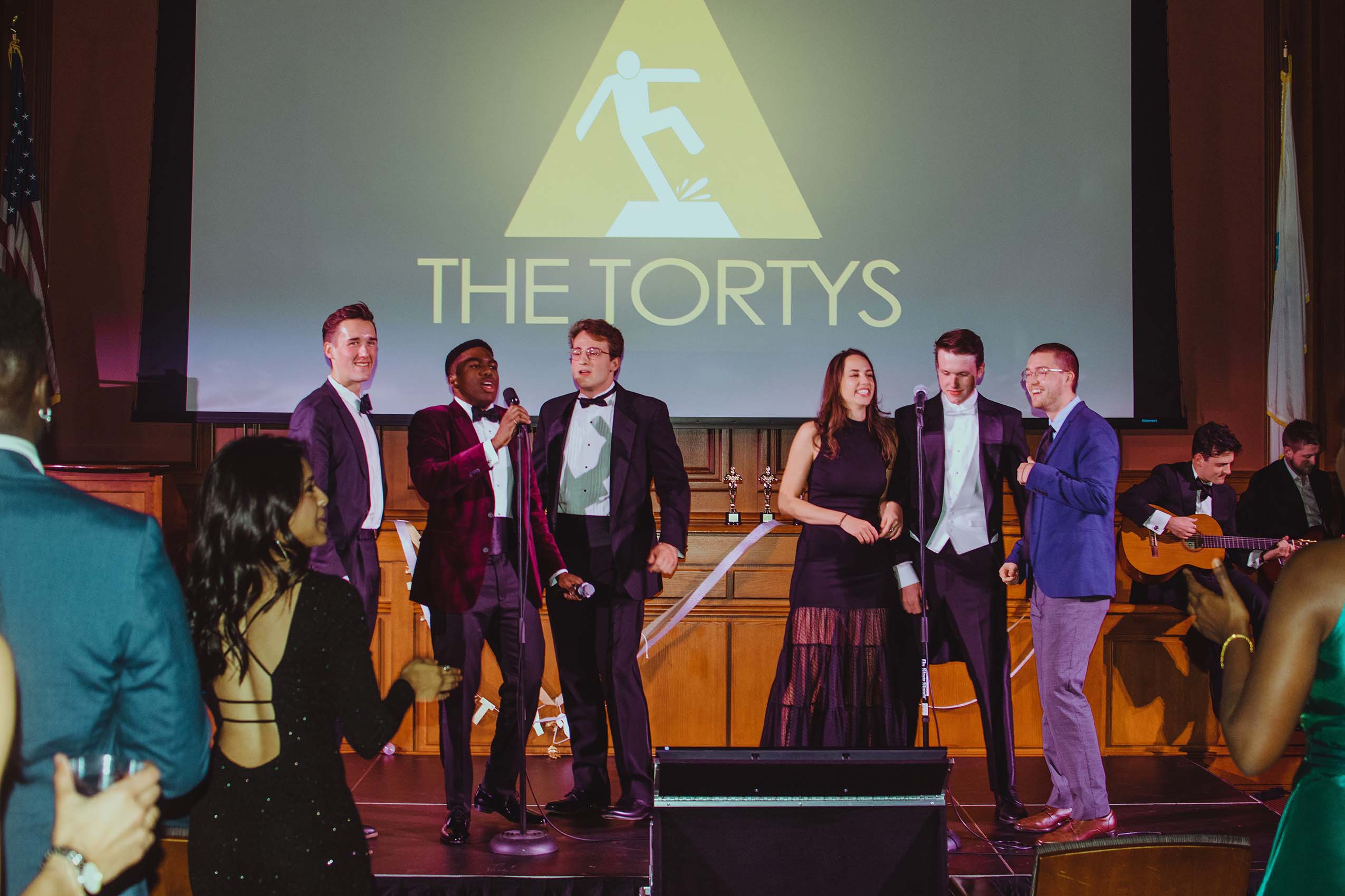Six studentes wearing formal attire standing on a stage, a video screen behind them reads: THE TORTYS.