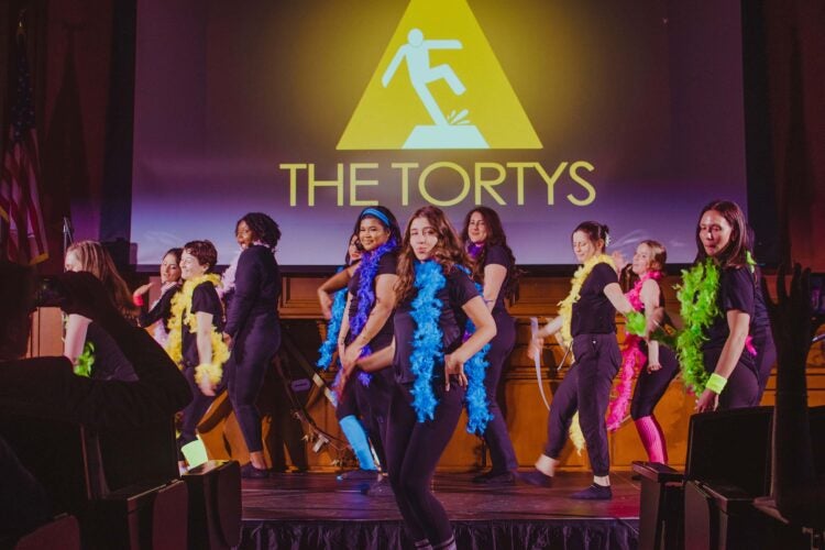 A group of women wearing all black and different colored boas on stage.