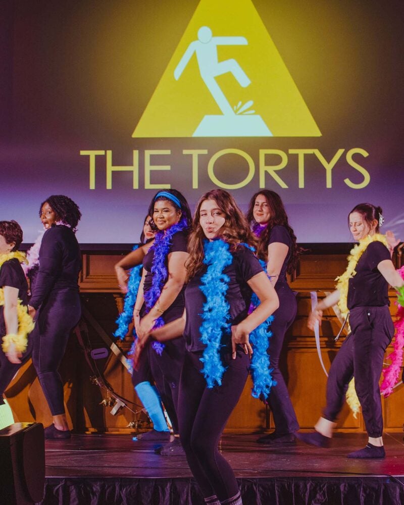 A group of women wearing all black and different colored boas on stage.