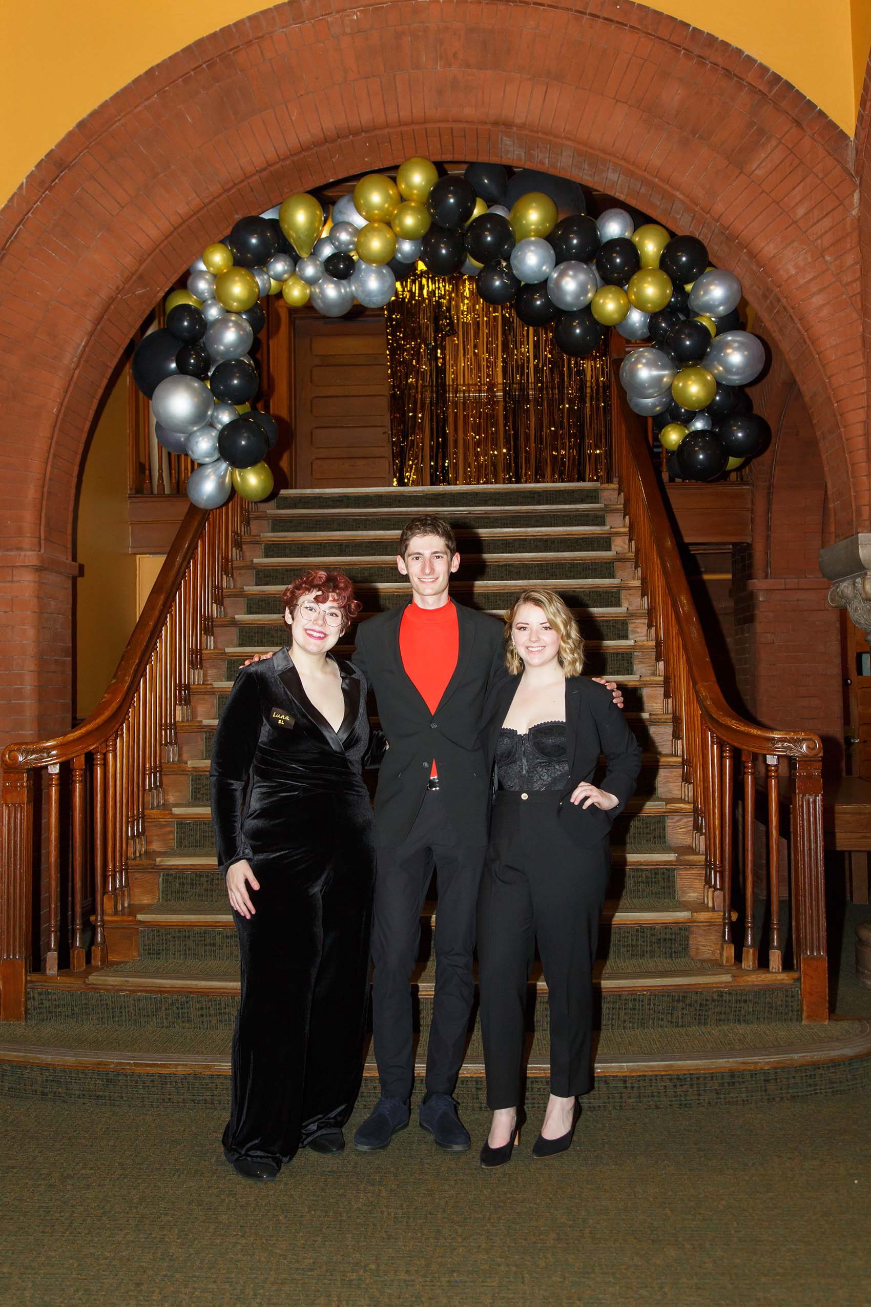 Two women and a man wearing formal attire pose under an arch of balloons.