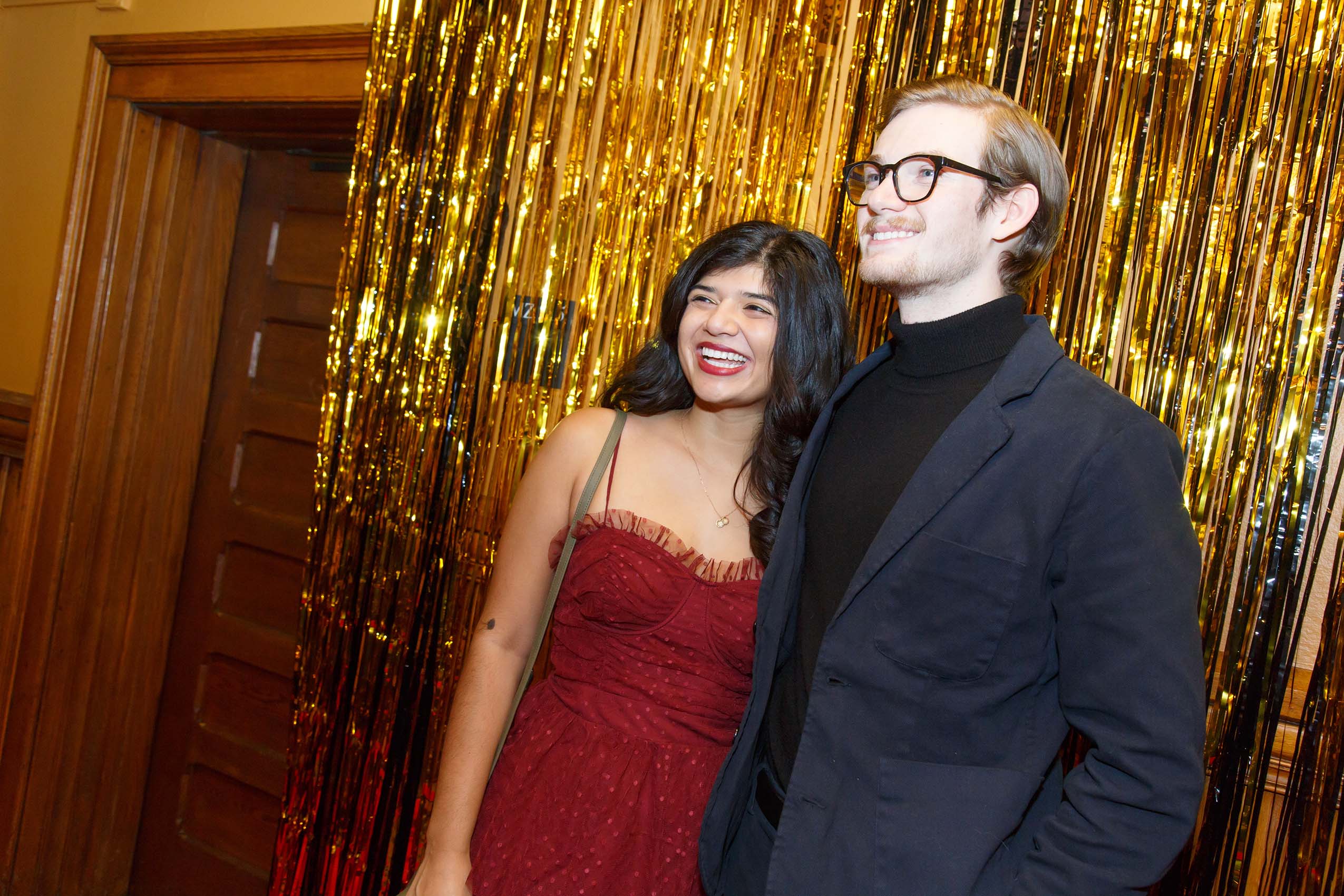 A couple wearing formal attire poses in front of a gold backdrop.