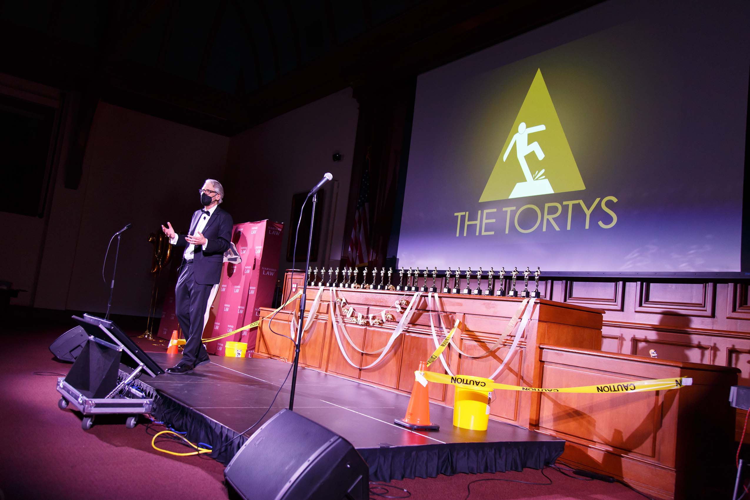 Professor Jon Hanson, wearing a face mask, stands on a stage. Behind him is a large screen that says: THE TORTYS and an image of a person falling.