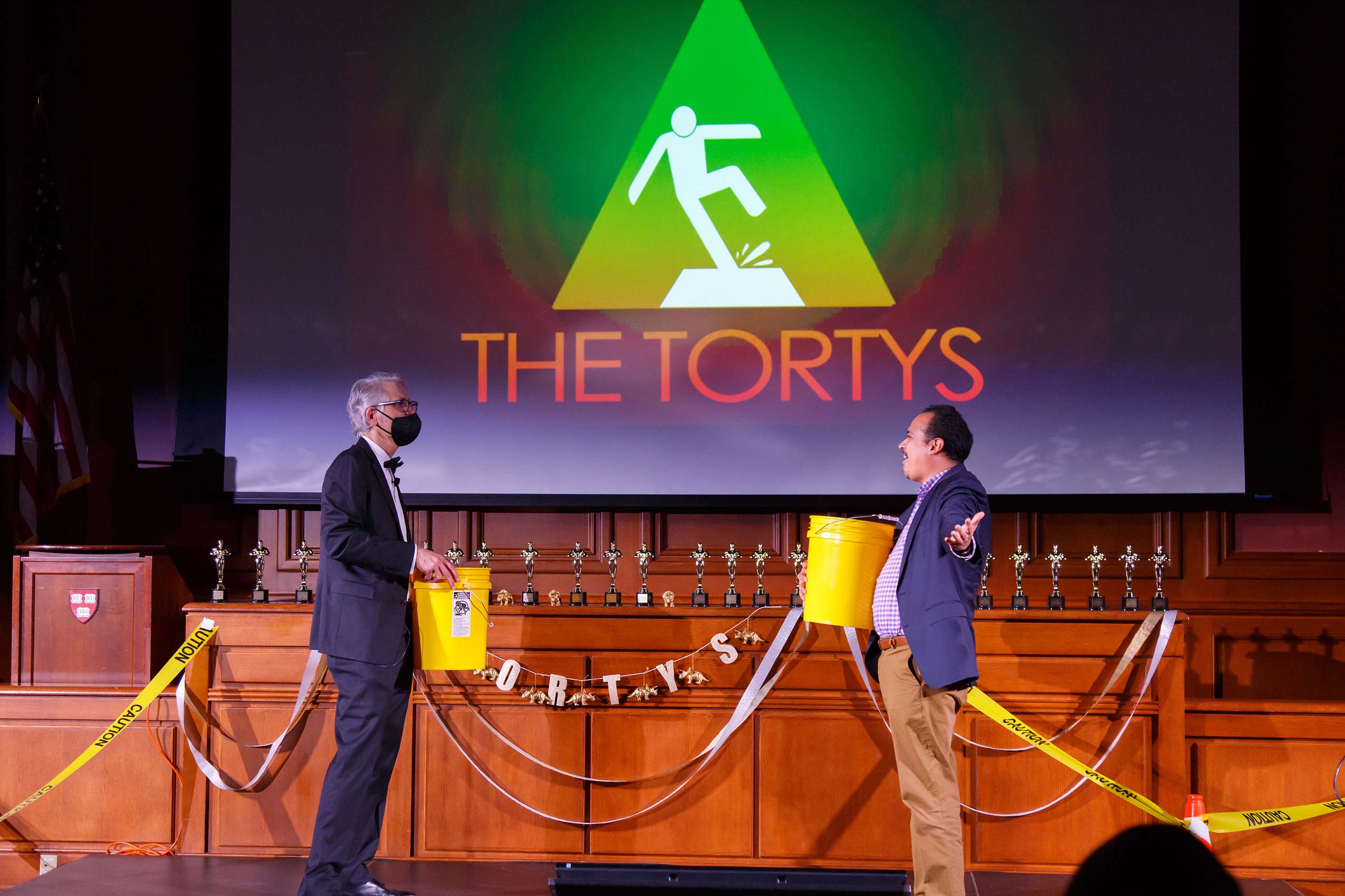 Professor Jon Hanson and a man wearing a blazer face each other on stage holding yellow buckets.