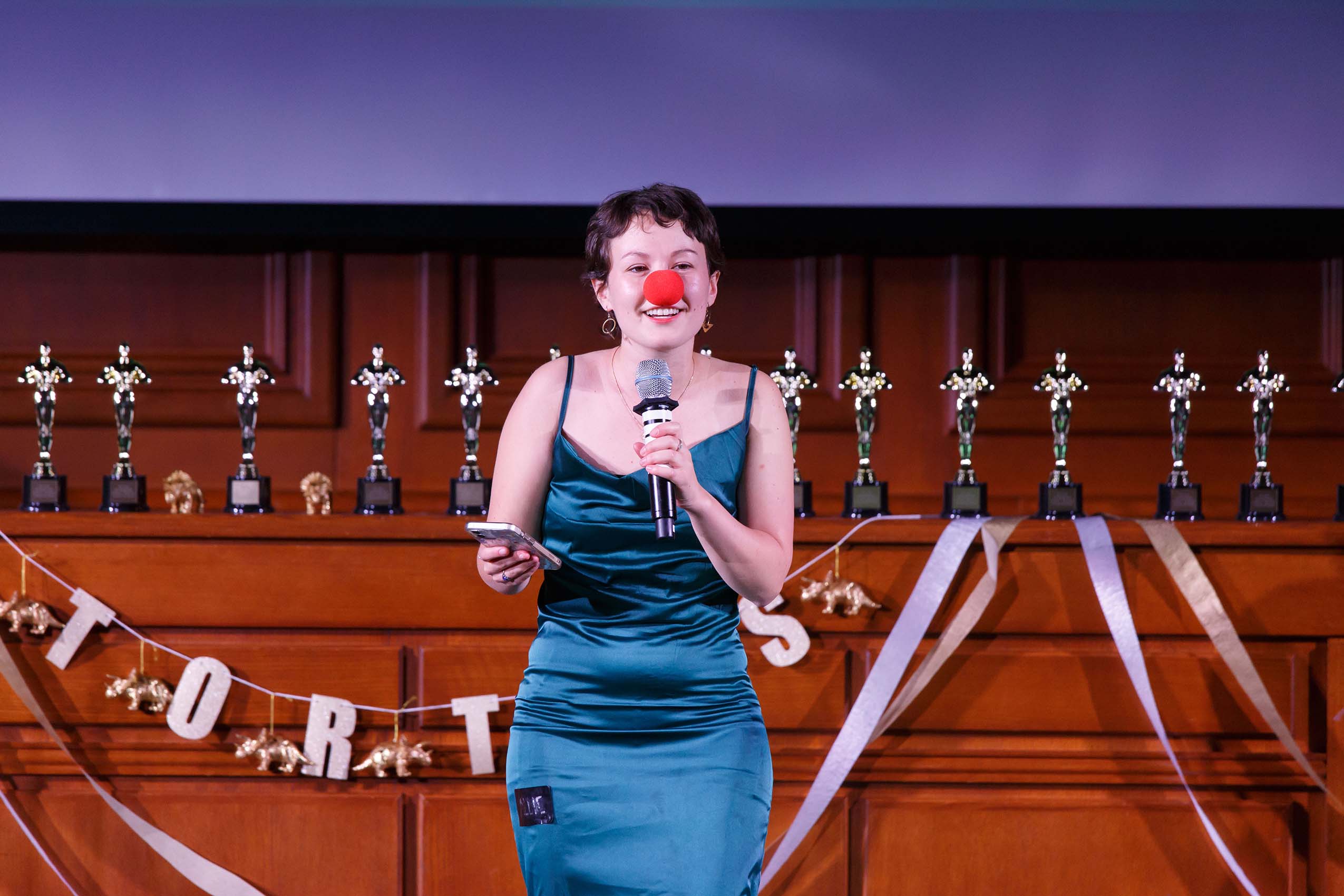 A woman in a teal dress and red nose standing on a stage with a microphone.