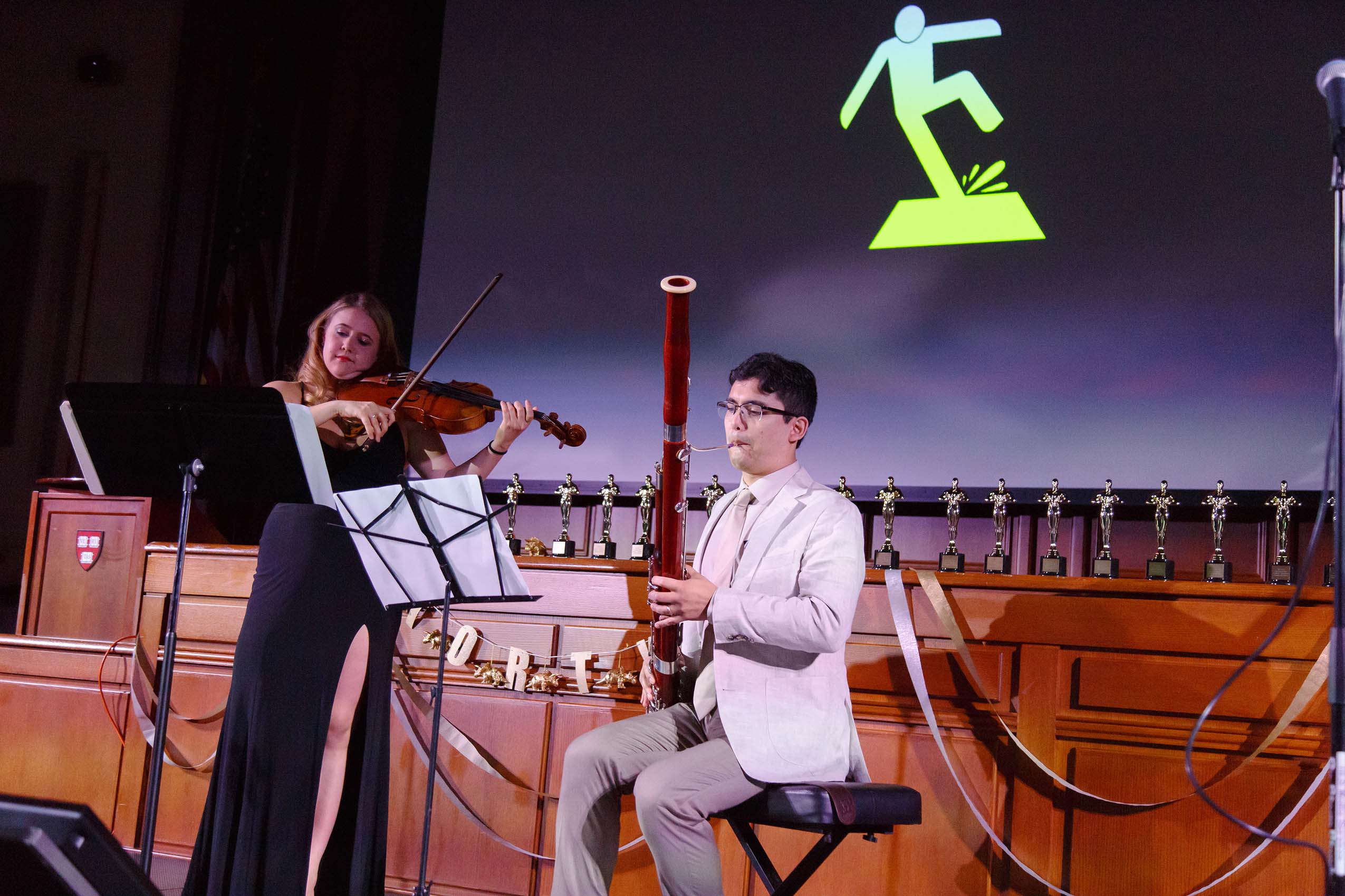 A woman in a formal gown stands, playing the violin. And man in a suit sits next to her, playing a bassoon..