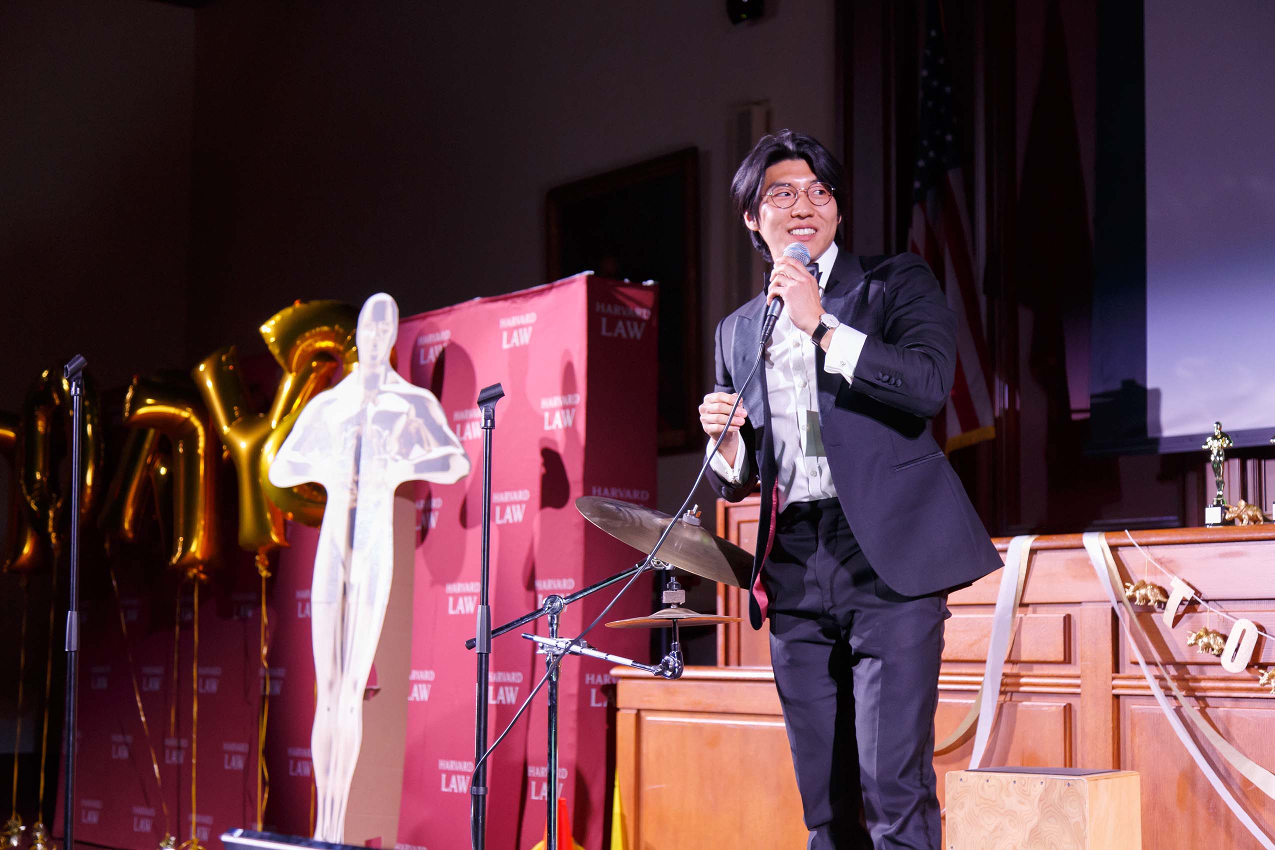 A student wearing a tuxedo stands on stage, holding a microphone.