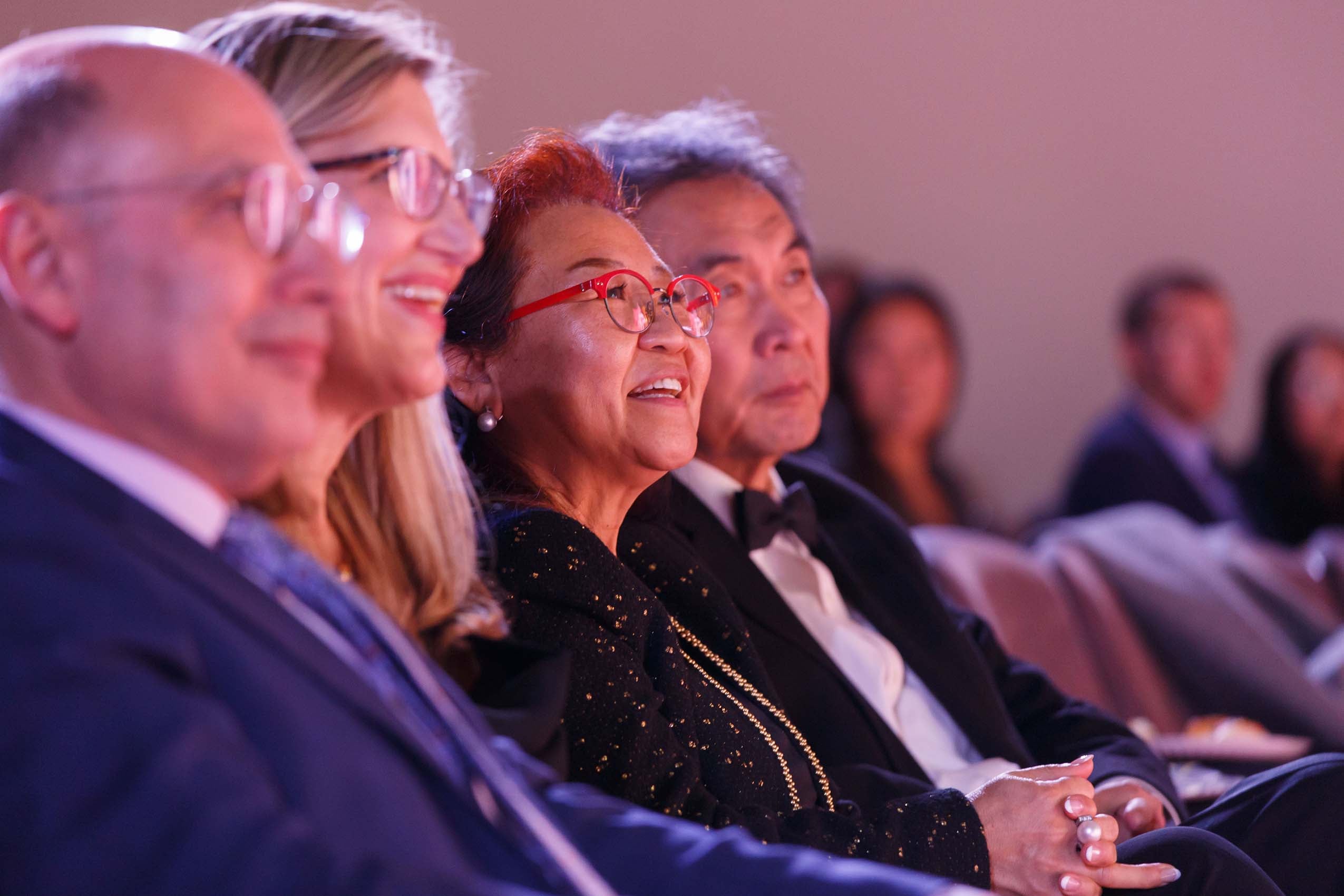 Four older adults in an audience, focus on a woman wearing red glasses smiling.
